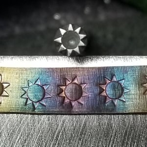 May include: Close-up of a metal stamping tool and a metal strip with stamped sun designs. The strip has a gradient of colors, including gold, blue, and purple. The sun stamps are evenly spaced and detailed.