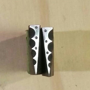 Puede incluir: Dos objetos metálicos con un acabado oscuro y un patrón decorativo de círculos y líneas curvas alternas. Los objetos están de pie, uno al lado del otro, sobre un fondo neutro.