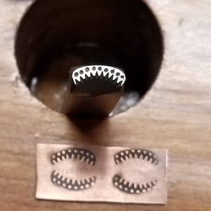May include: Close-up of a metal stamp with a decorative edge design. The stamp is positioned near a wooden surface with a circular hole. Below, a copper plate displays two stamped impressions of the design.