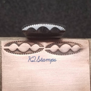 Puede incluir: Un sello negro con un diseño festoneado blanco sobre una placa de metal color cobre. La placa tiene dos impresiones ovaladas con un diseño similar. El texto "K2 Stamps" está escrito en azul.