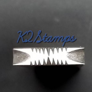 Puede incluir: Un sello metálico rectangular con un patrón en zigzag y el texto "K2 Stamps" en cursiva azul. El sello tiene una superficie texturizada y un diseño en zigzag blanco.