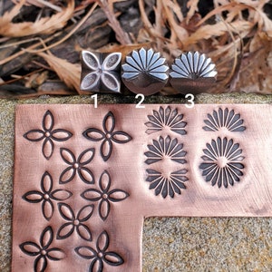 Puede incluir: Sellos metálicos con diseños florales y de rayos de sol, junto con una lámina de cobre con impresiones estampadas. Los sellos se utilizan para crear diseños. La lámina de cobre muestra los patrones estampados. Los sellos están numerados 1, 2 y 3.