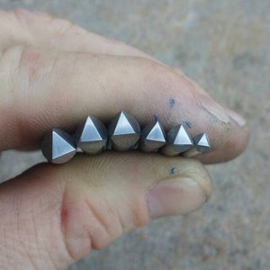 May include: A set of six silver-colored, geometric metal stamps in varying sizes, each with a pyramidal shape. The stamps are held in a hand, showcasing their polished surfaces and sharp edges. The background is a neutral, textured surface.