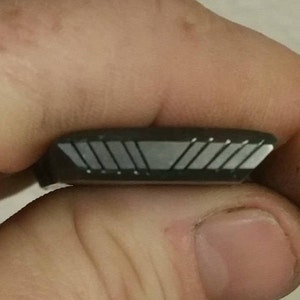 Puede incluir: Un pequeño objeto rectangular gris oscuro con una serie de líneas grises claras en ángulo en un lado. El objeto se sostiene entre dos dedos, mostrando su diseño geométrico y acentos metálicos.