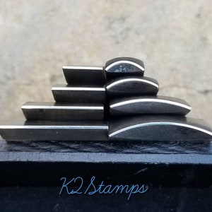 Könnte beinhalten: Eine Ansammlung dunkelgrauer Metallstempel mit silberner Kante, pyramidenförmig angeordnet. Die Stempel sind auf einer schwarzen Oberfläche angeordnet, mit dem Text "K2 Stamps" in Hellblau unten.