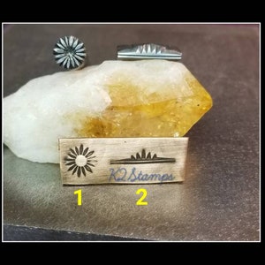 May include: Metal stamps with floral and geometric designs. The image shows a sunflower stamp, a geometric stamp, and a stamp with the text "K2 Stamps". The stamps are on a stone surface.
