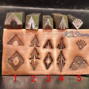 Puede incluir: Un conjunto de sellos metálicos con diseños geométricos, incluyendo rayos de sol y árboles estilizados. Los sellos están dispuestos sobre un bloque color cobre, con el texto "K2 Stamps" visible. Herramientas para crear impresiones.
