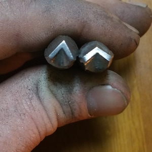 May include: Two small, silver-colored metal objects, each with a V-shaped design, are held in a hand. The objects appear to be tools or components, possibly for jewelry making or metalworking. The hand is covered in a fine, dark residue.