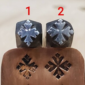 May include: Two metal stamps with a snowflake design are shown. The stamps are dark grey with a silver snowflake pattern. Below the stamps is a copper-coloured metal piece with the same snowflake design stamped into it.