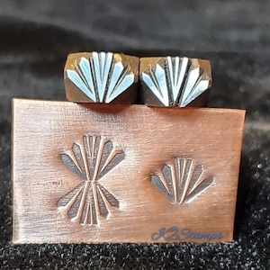 May include: Two metal stamps with a fan-like design sit on a copper plate with matching impressions. The plate has the text "K2 Stamps" in the lower right corner. The stamps and plate are on a dark background.