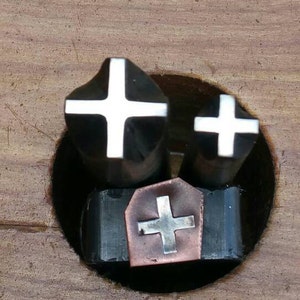 May include: Three metal cross stamps with white cross designs. The stamps are black with white crosses. One stamp is on a copper-colored base. The stamps are set in a wooden surface.