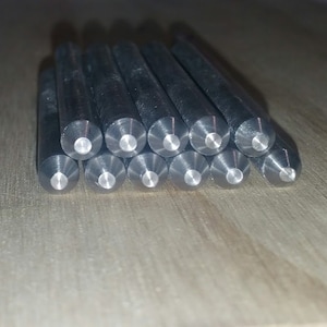 May include: A collection of cylindrical metal punches with a conical tip and a flat, circular end. The punches are arranged in a slightly angled, stacked formation, showcasing their precision and design. The metal has a dark, reflective finish.