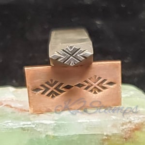 May include: A metal stamp with a geometric snowflake design. The stamp is silver and is positioned above a copper-colored metal plate with the same snowflake pattern stamped into it. The plate has the text "K2 Stamps" in the lower right corner.