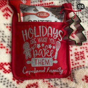 Pode incluir: Uma luva de forno vermelha com o texto prateado brilhante "HOLIDAYS ARE WHAT YOU BAKE THEM Copeland Family". Contém uma mistura de crumble Betty Crocker, uma colher de pau e um cortador de biscoitos em forma de árvore de Natal.