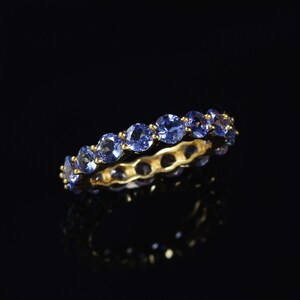 May include: A gold band ring with a row of blue gemstones. The ring is on a black background.