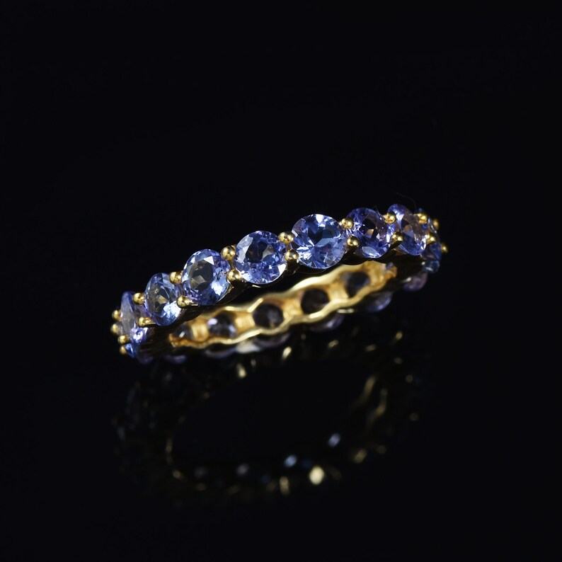 May include: A gold band ring with a row of blue gemstones. The ring is on a black background.