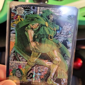 May include: A trading card featuring a comic book illustration of Doctor Doom in a green hooded cloak and armor. The card has the text "Your Turn +1000" at the bottom. The background shows comic book panels.