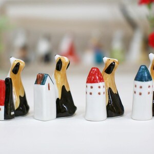 May include: A set of seven ceramic figurines, including houses and dogs. The houses are white with red, blue, and green accents. The dogs are brown and black with white accents. The figurines are all facing forward and are arranged in a row.