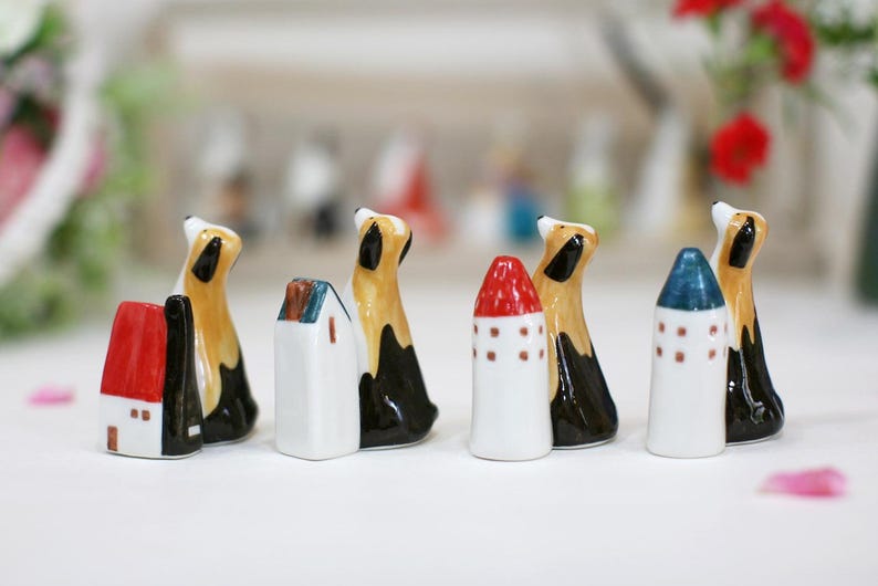 May include: A set of seven ceramic figurines, including houses and dogs. The houses are white with red, blue, and green accents. The dogs are brown and black with white accents. The figurines are all facing forward and are arranged in a row.