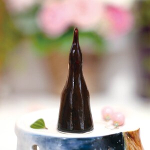 May include: A dark brown ceramic bottle stopper shaped like a tall, narrow bottle. The stopper is sitting on a white ceramic base with a blue and white design.