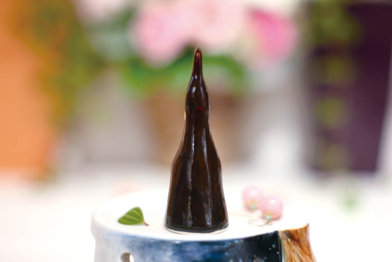 May include: A dark brown ceramic bottle stopper shaped like a tall, narrow bottle. The stopper is sitting on a white ceramic base with a blue and white design.
