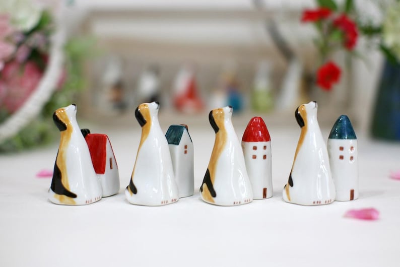 May include: A set of eight ceramic figurines, each depicting a dog with a house on its back. The houses are painted in various colors, including red, blue, and green. The dogs are all looking up, and their tails are wagging.
