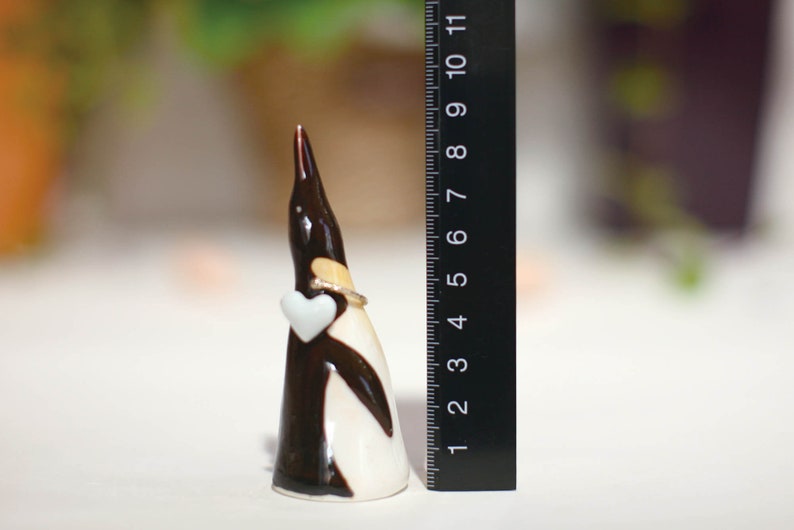 May include: A ceramic penguin figurine holding a white heart. The penguin is black and white with a gold ring around its neck. The figurine is approximately 5 inches tall.
