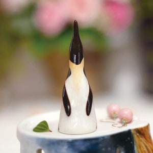 May include: A small ceramic penguin figurine with black and white markings. The penguin is standing on a white pedestal with a green leaf.