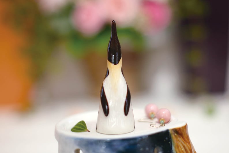 May include: A small ceramic penguin figurine with black and white markings. The penguin is standing on a white pedestal with a green leaf.