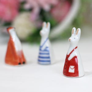 Puede incluir: Tres figuritas de cerámica de animales con ropa. Un conejo blanco con un vestido rojo, un conejo blanco con un vestido a rayas azul y blanco y un zorro con un vestido naranja.