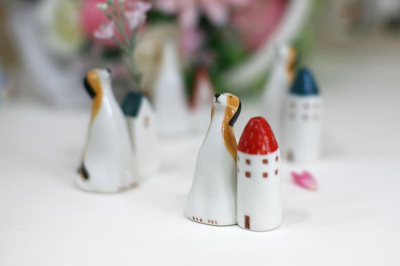 May include: A set of ceramic figurines depicting a dog and a house with a red roof. The figurines are painted in white, brown, red, and blue.