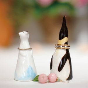 May include: White ceramic penguin and polar bear ring holders with gold bands around their necks. The penguin has a black and white design and the polar bear has a blue and white design. Two pink and green decorative beads are in front of the holders.