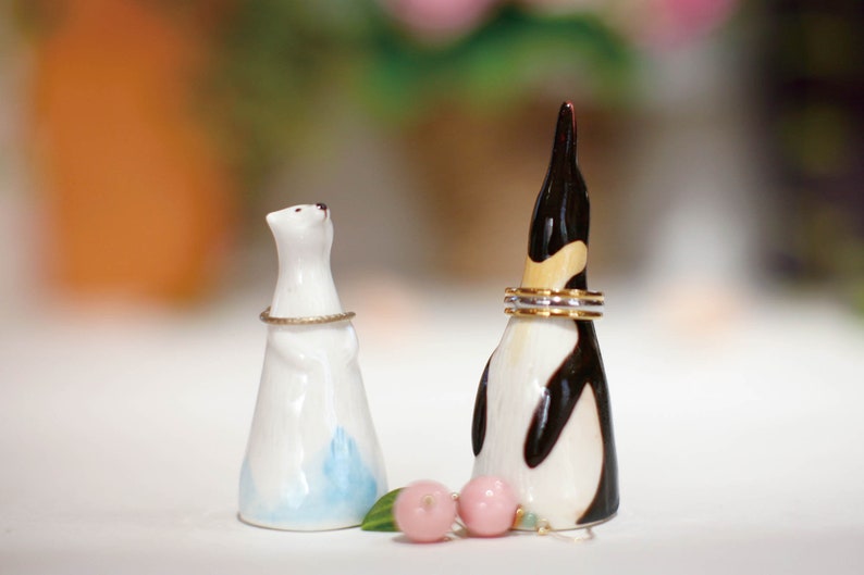 May include: White ceramic penguin and polar bear ring holders with gold bands around their necks. The penguin has a black and white design and the polar bear has a blue and white design. Two pink and green decorative beads are in front of the holders.