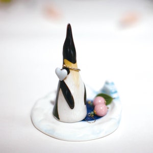 May include: A ceramic penguin ring holder with a white heart and a gold ring. The penguin is standing on a white and blue ceramic base with two pink beads and a green leaf.
