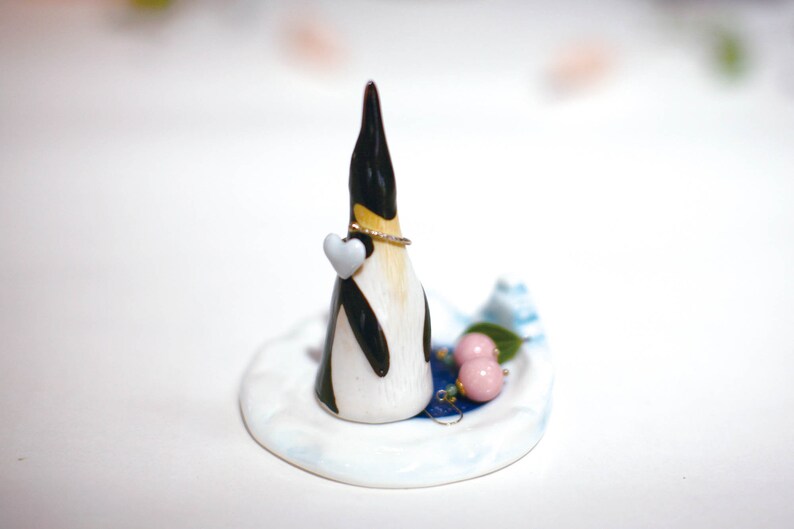May include: A ceramic penguin ring holder with a white heart and a gold ring. The penguin is standing on a white and blue ceramic base with two pink beads and a green leaf.