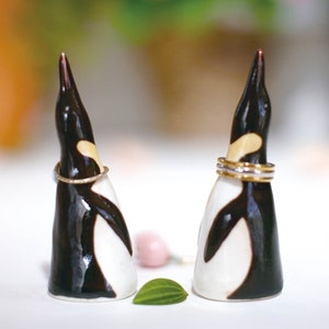 May include: Two black and white ceramic penguin ring holders with gold bands around their necks. The penguins are facing each other and are on a white surface.