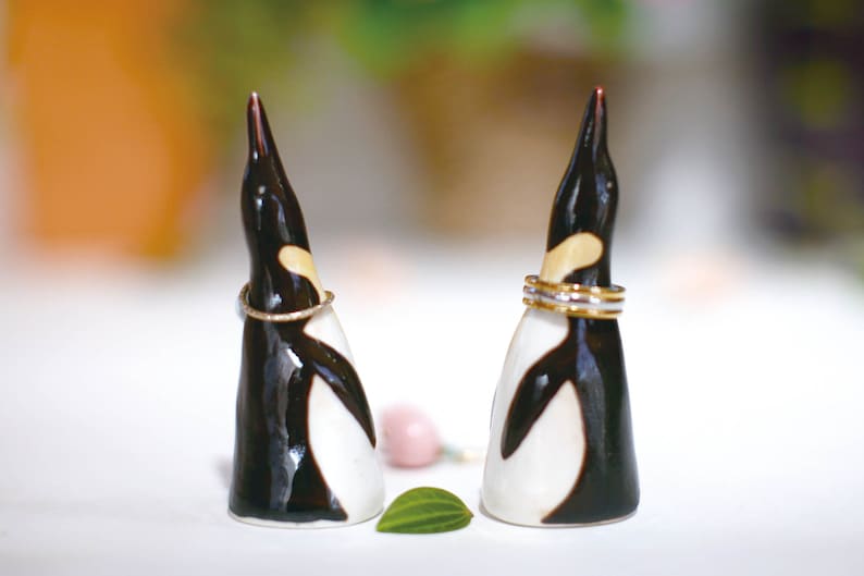 May include: Two black and white ceramic penguin ring holders with gold bands around their necks. The penguins are facing each other and are on a white surface.