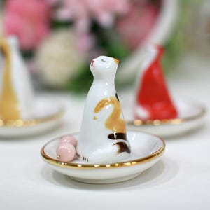 May include: A white ceramic cat ornament with calico markings, on a small, round dish with a gold rim. Two pink beads are near the cat. Other similar ornaments are in the background.