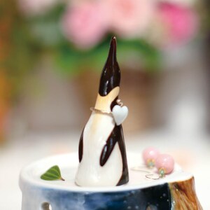 May include: A ceramic penguin figurine with a white heart in its beak, holding a ring on a small wooden stand with a blue and white design.