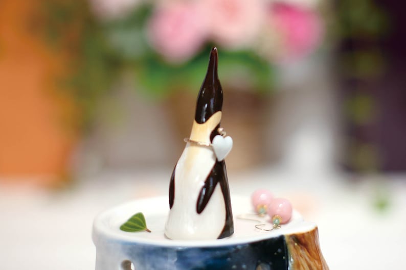 May include: A ceramic penguin figurine with a white heart in its beak, holding a ring on a small wooden stand with a blue and white design.