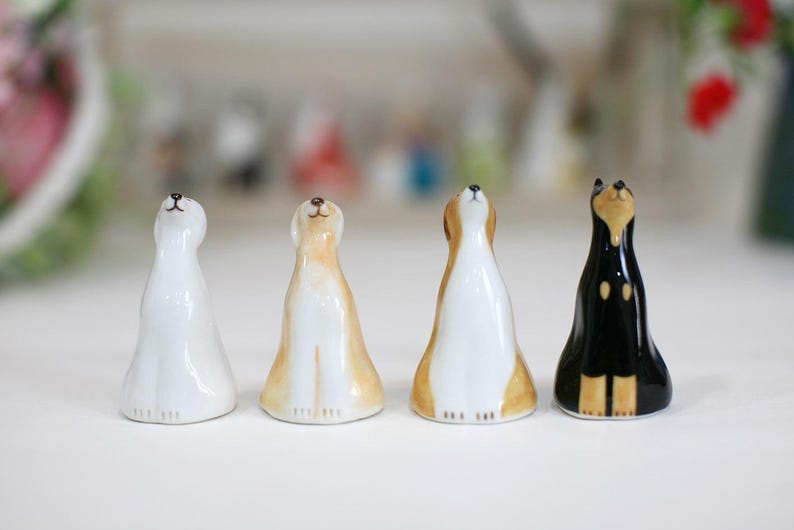 May include: Four ceramic figurines of dogs, each with a different color and design. The dogs are all looking up, and they are standing on a white surface.