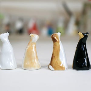 May include: Four ceramic dog figurines in different colors and poses. The dogs are all looking up, with their heads tilted back. The figurines are made of white, brown, tan, and black ceramic.