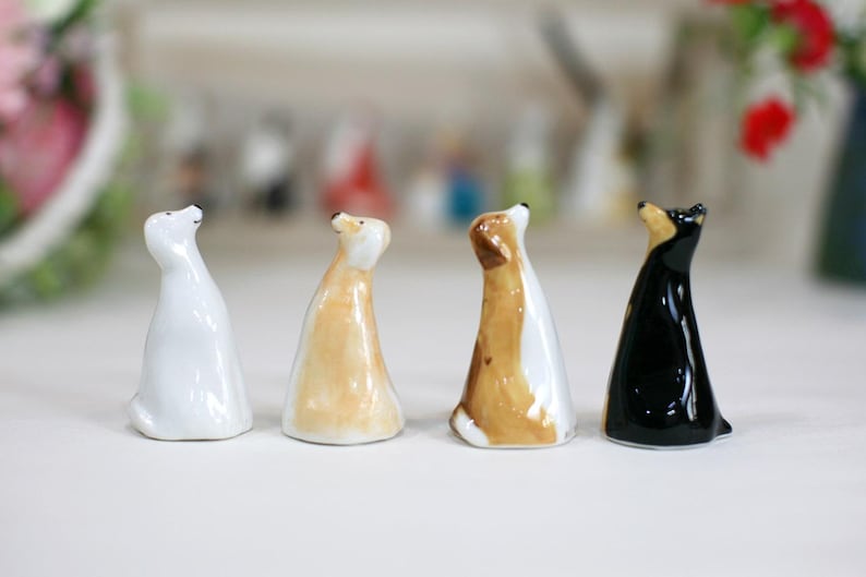 May include: Four ceramic dog figurines in different colors and poses. The dogs are all looking up, with their heads tilted back. The figurines are made of white, brown, tan, and black ceramic.