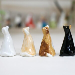 May include: Four ceramic cat figurines in different colors: white, tan, brown and black. The cats are all sitting upright with their heads tilted up.