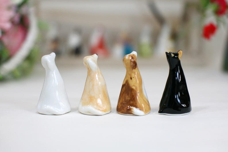 May include: Four ceramic cat figurines in different colors: white, tan, brown and black. The cats are all sitting upright with their heads tilted up.