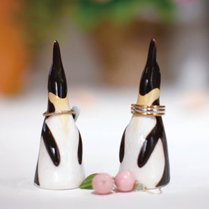 May include: Two ceramic penguin figurines with black and white markings, used as ring holders. The penguin on the left has a single gold ring around its neck, while the penguin on the right has three gold rings around its neck.  There are two pink and green earrings in front of the penguins.