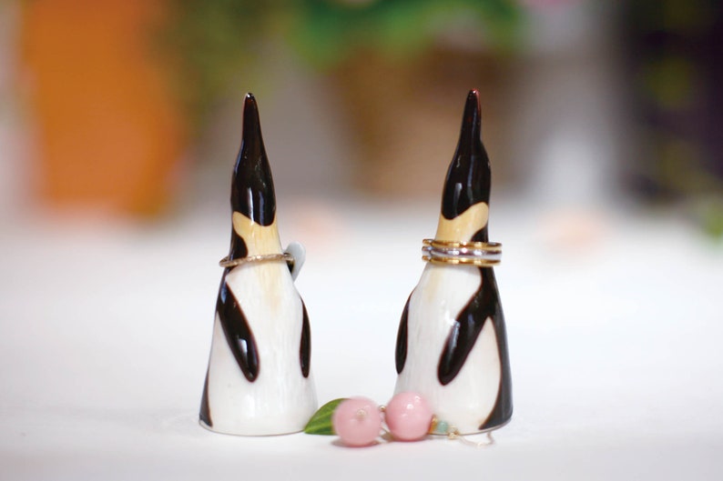 May include: Two ceramic penguin figurines with black and white markings, used as ring holders. The penguin on the left has a single gold ring around its neck, while the penguin on the right has three gold rings around its neck.  There are two pink and green earrings in front of the penguins.