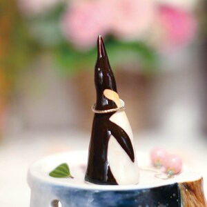 May include: A ceramic penguin ring holder with a black and white design. The penguin is holding a gold ring. The ring holder is sitting on a white and blue ceramic base.