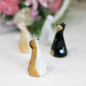 May include: Three ceramic dog figurines, each with a different color and design. The dog on the left is white with brown spots, the dog in the middle is white with brown and black spots, and the dog on the right is black.