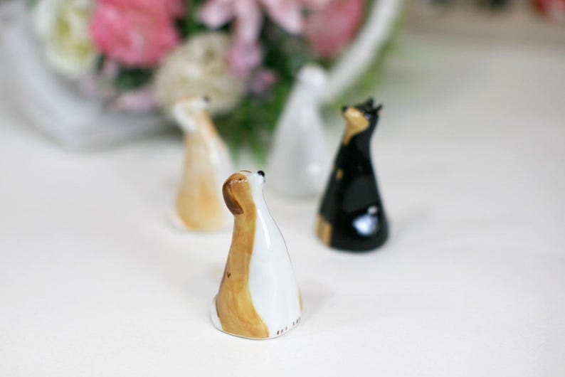 May include: Three ceramic dog figurines, each with a different color and design. The dog on the left is white with brown spots, the dog in the middle is white with brown and black spots, and the dog on the right is black.